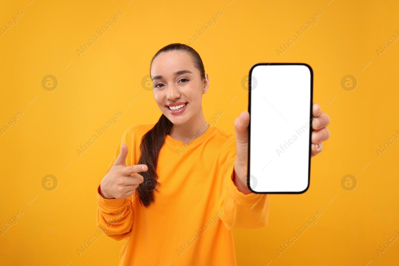 Photo of Young woman showing smartphone in hand and pointing at it on yellow background