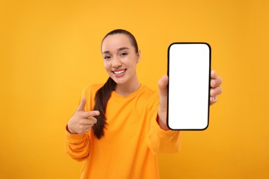 Photo of Young woman showing smartphone in hand and pointing at it on yellow background