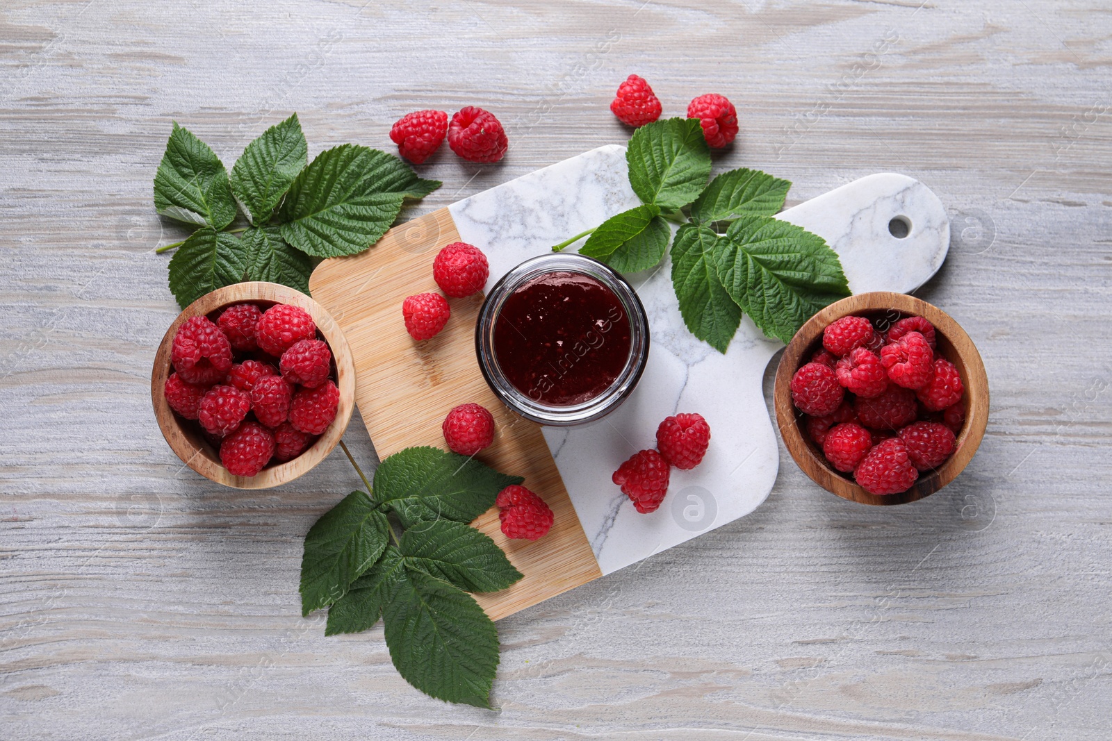 Photo of Delicious raspberry jam, fresh berries and green leaves on light wooden table, flat lay