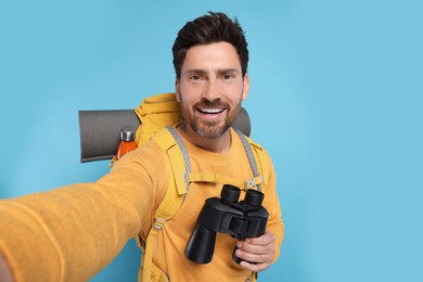 Photo of Happy man with backpack and binoculars taking selfie on light blue background. Active tourism