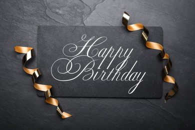 Photo of Slate board with greeting HAPPY BIRTHDAY and serpentine streamers on black background, top view