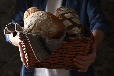 Photo of Man holding wicker basket with different types of bread on dark background, closeup