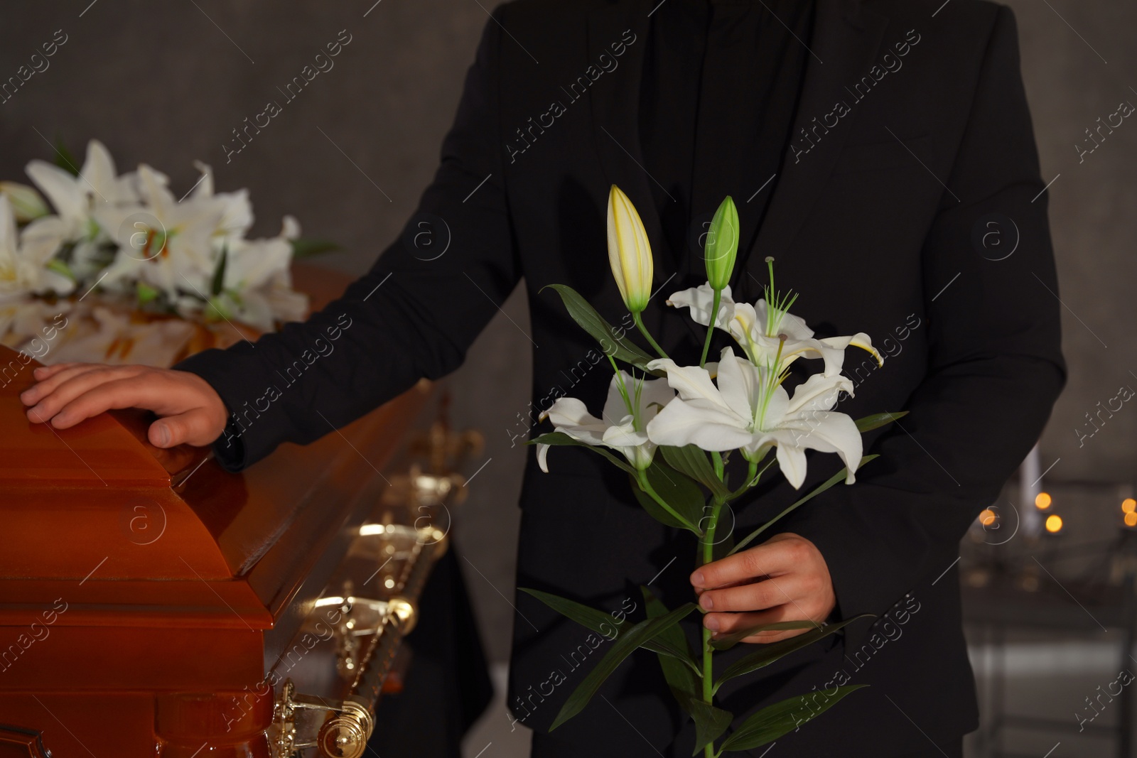 Photo of Young man with white lilies near casket in funeral home, closeup