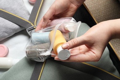 Woman putting small bottle with personal care product into plastic bag near suitcase, closeup. Cosmetic travel kit