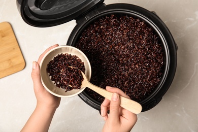 Woman putting delicious brown rice into bowl from multi cooker at table, top view