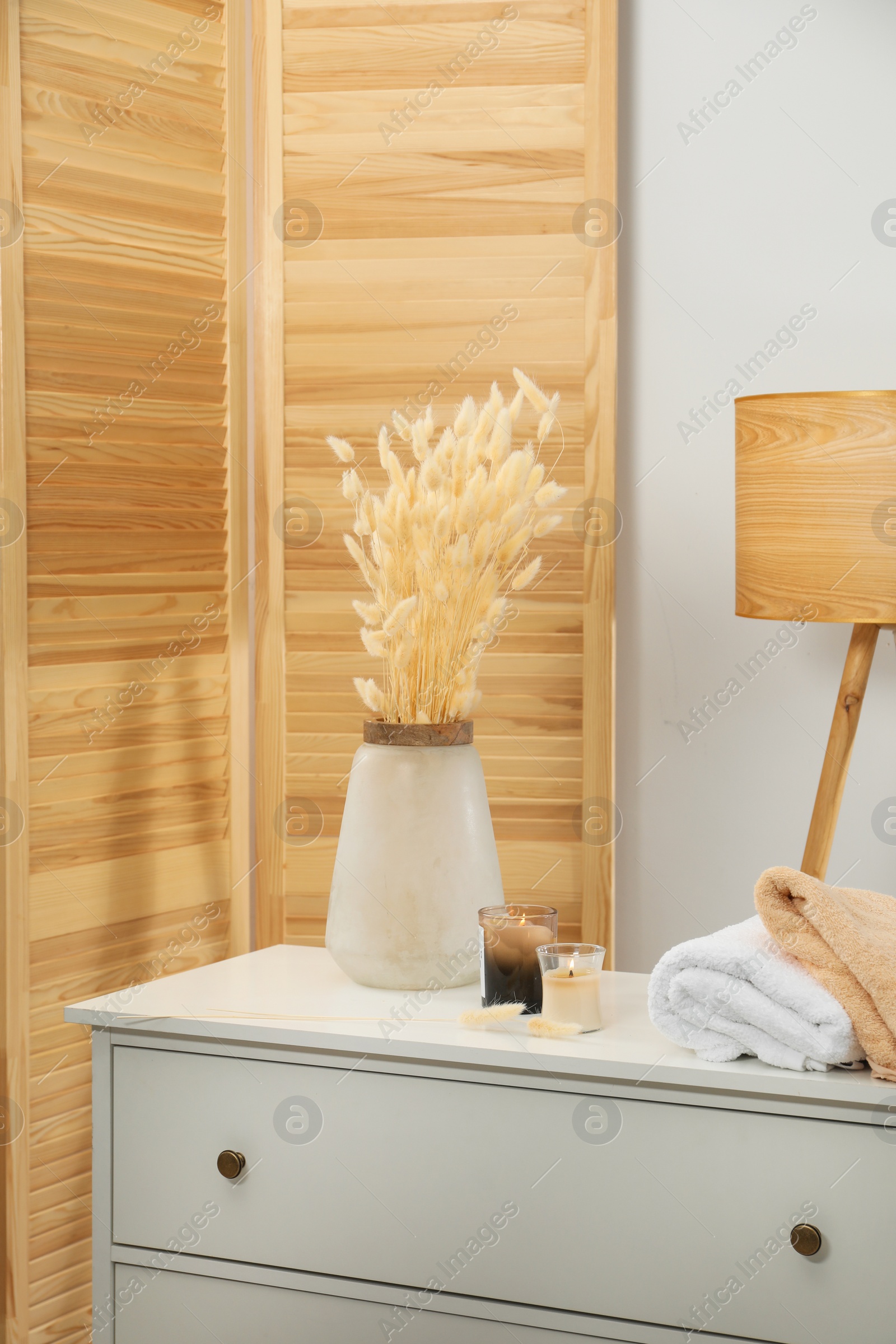 Photo of Vase with dried flowers, stacked towels and candles on chest of drawers in room. Interior design