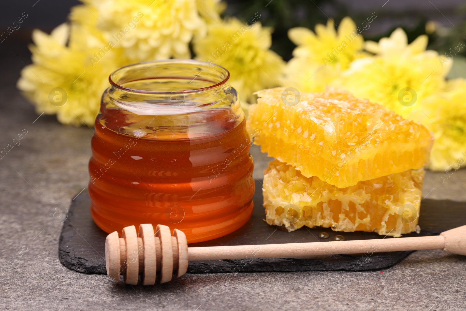 Photo of Sweet golden honey in jar, dipper and pieces of honeycomb on grey textured table, closeup