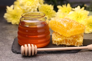 Sweet golden honey in jar, dipper and pieces of honeycomb on grey textured table, closeup