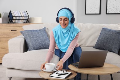Muslim woman with cup of drink using laptop at wooden table in room