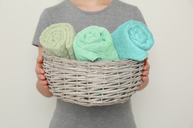 Photo of Woman holding wicker basket with rolled soft terry towels on light background, closeup