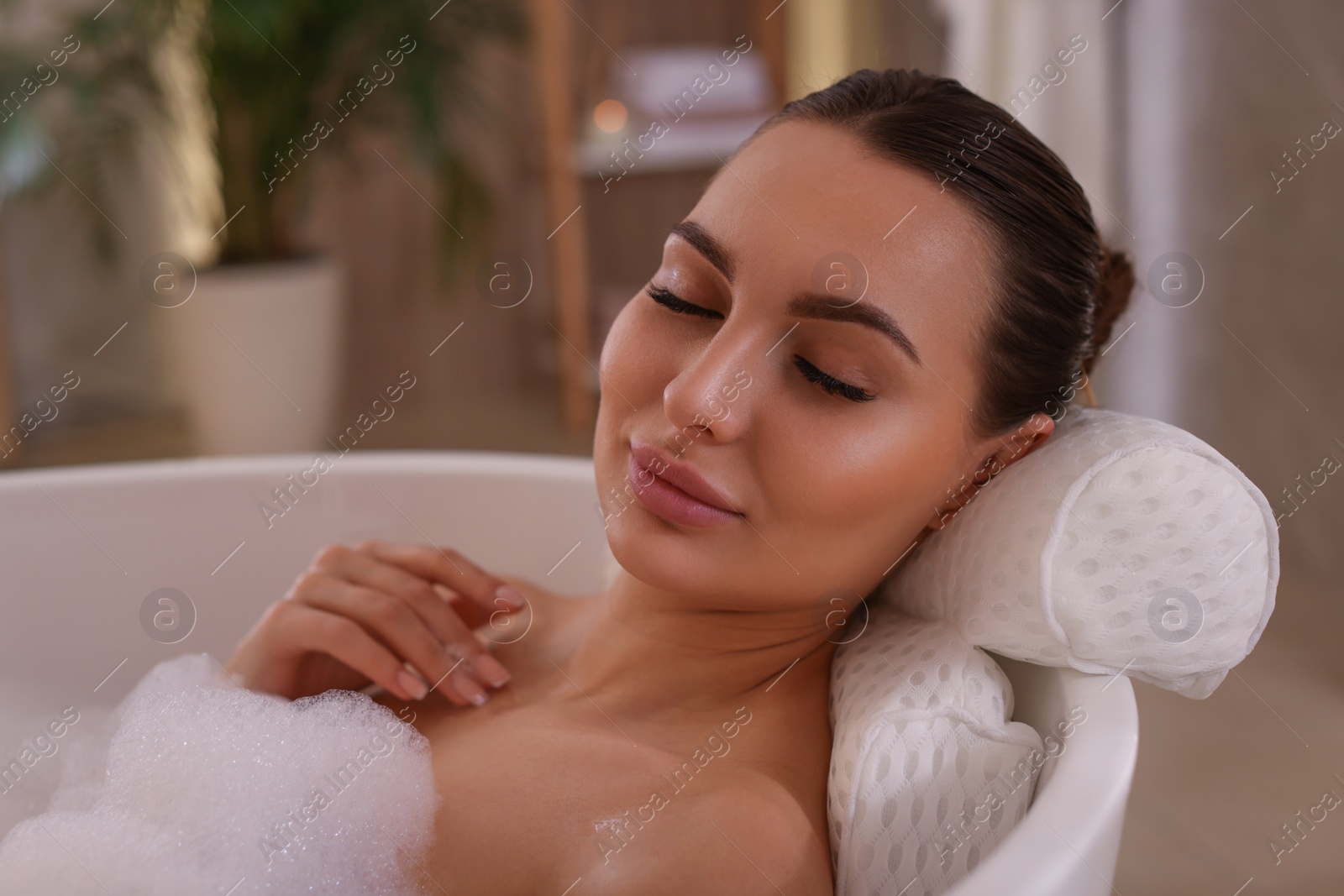 Photo of Young woman using pillow while enjoying bubble bath indoors