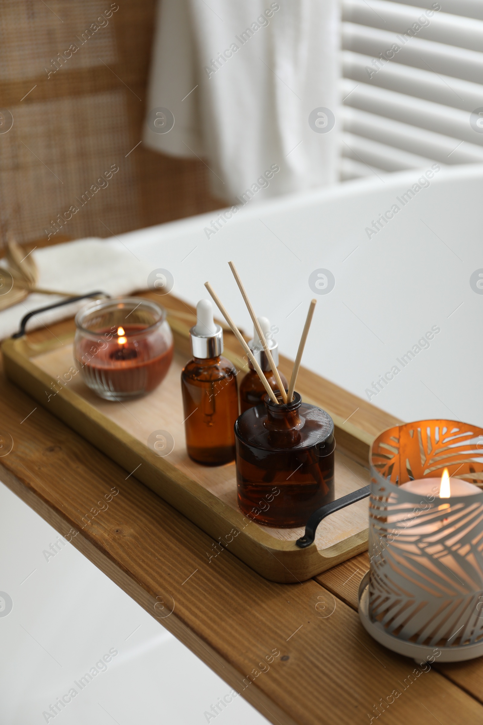 Photo of Wooden tray with cosmetic products, reed air freshener and burning candles on bath tub in bathroom