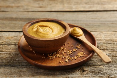 Photo of Spoon and bowl of tasty mustard sauce with dry seeds on wooden table
