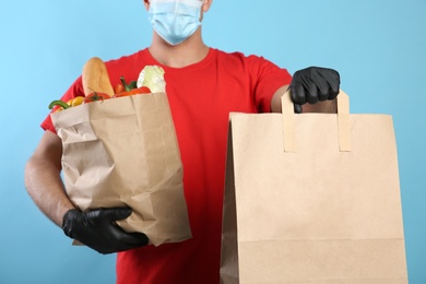 Photo of Courier in medical mask holding paper bags with takeaway food on light blue background, closeup. Delivery service during quarantine due to Covid-19 outbreak