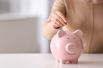 Photo of Woman putting coin into piggy bank at table indoors, closeup. Space for text