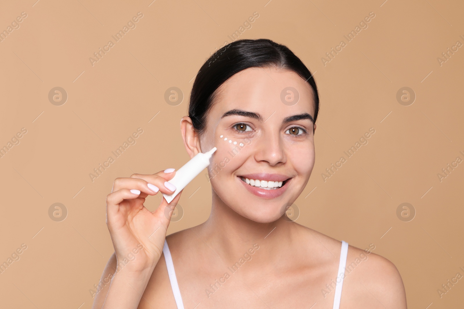 Photo of Beautiful young woman applying cream on skin under eye against beige background