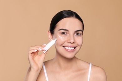 Photo of Beautiful young woman applying cream on skin under eye against beige background