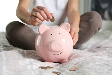 Photo of Woman putting money into piggy bank on bed at home, closeup