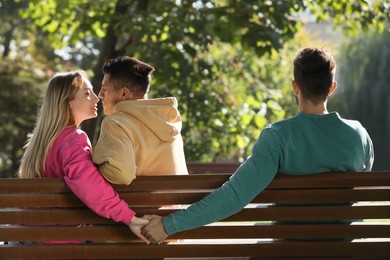 Photo of Woman holding hands with another man behind her boyfriend's back on bench in park. Love triangle