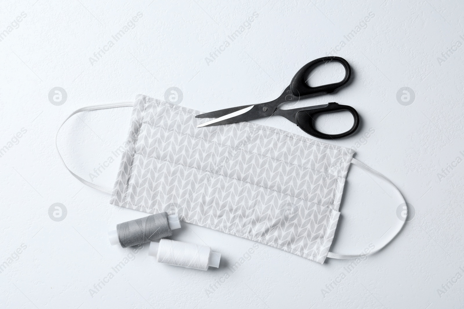 Photo of Homemade protective mask, threads and scissors on white background, flat lay. Sewing idea