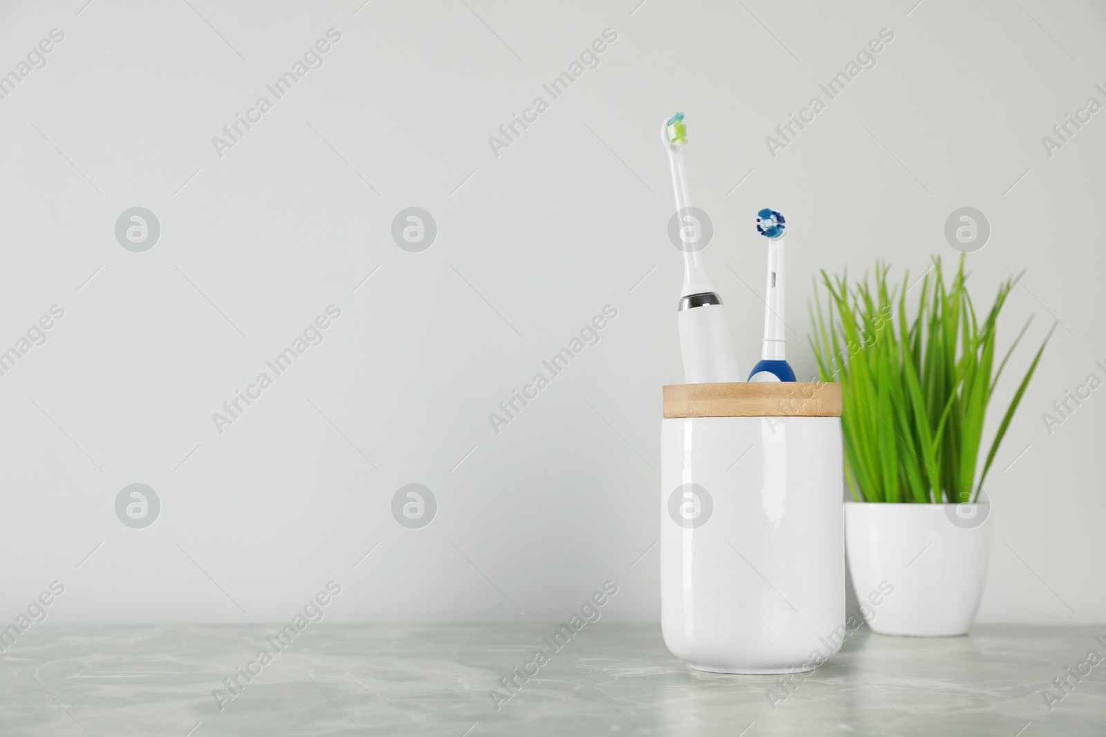 Photo of Electric toothbrushes in holder and green houseplant on light grey marble table near white wall. Space for text