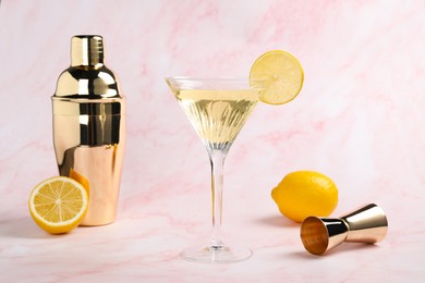 Martini glass of refreshing cocktail with lemon slice and fresh fruits near shaker on pink marble background