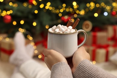 Photo of Woman holding cup of delicious cocoa with marshmallows near Christmas tree at home, closeup