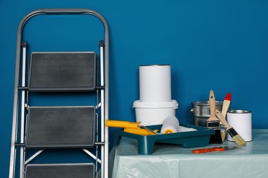 Photo of Metallic folding ladder and painting tools on table near blue wall