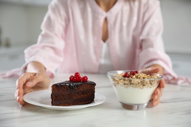Photo of Woman choosing between yogurt with granola and cake at white table in kitchen, closeup