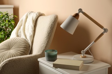 Photo of Stylish modern desk lamp, book and candle on white cabinet near soft armchair indoors