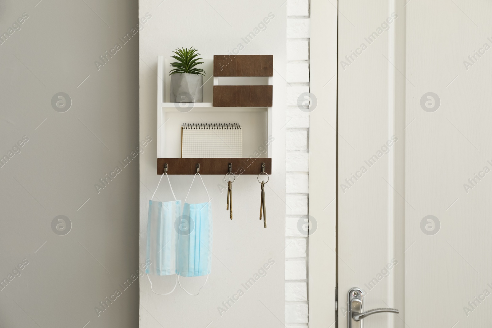 Photo of Hanger for keys with protective masks, houseplant and notebook on shelves near door in hallway