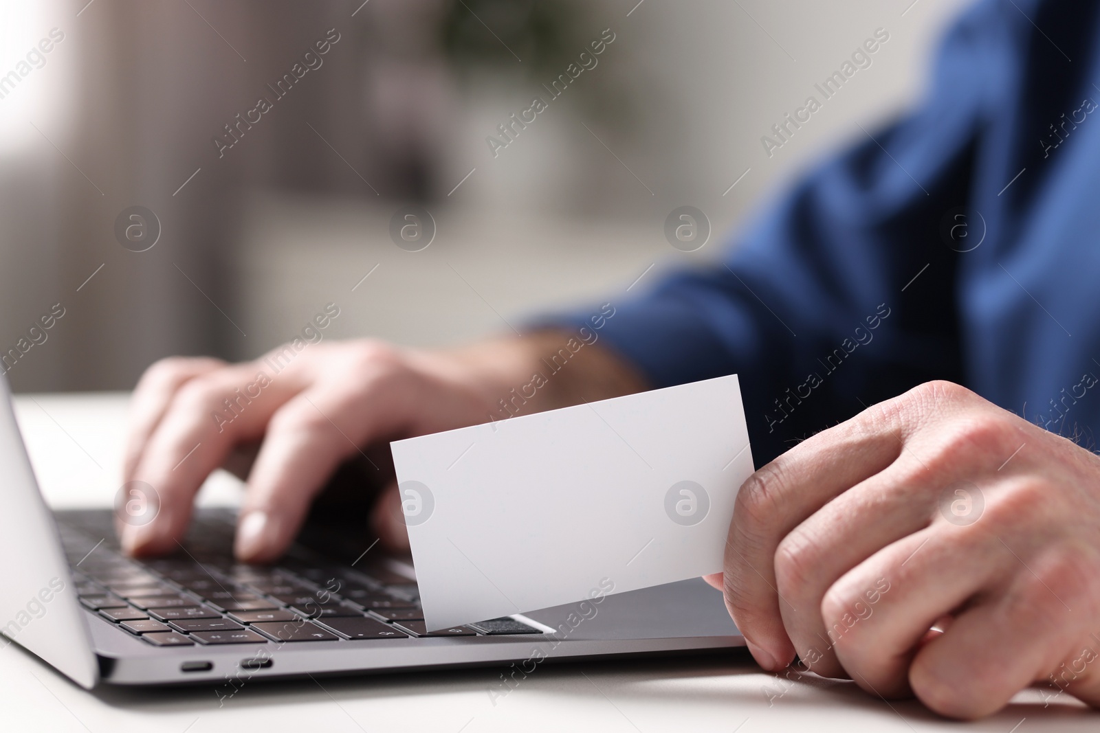 Photo of Man with laptop holding blank business card at white table indoors, closeup