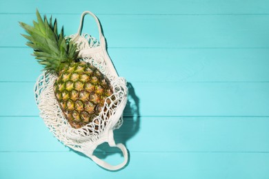 Photo of Whole ripe pineapple in mesh bag on light blue wooden table, top view. Space for text