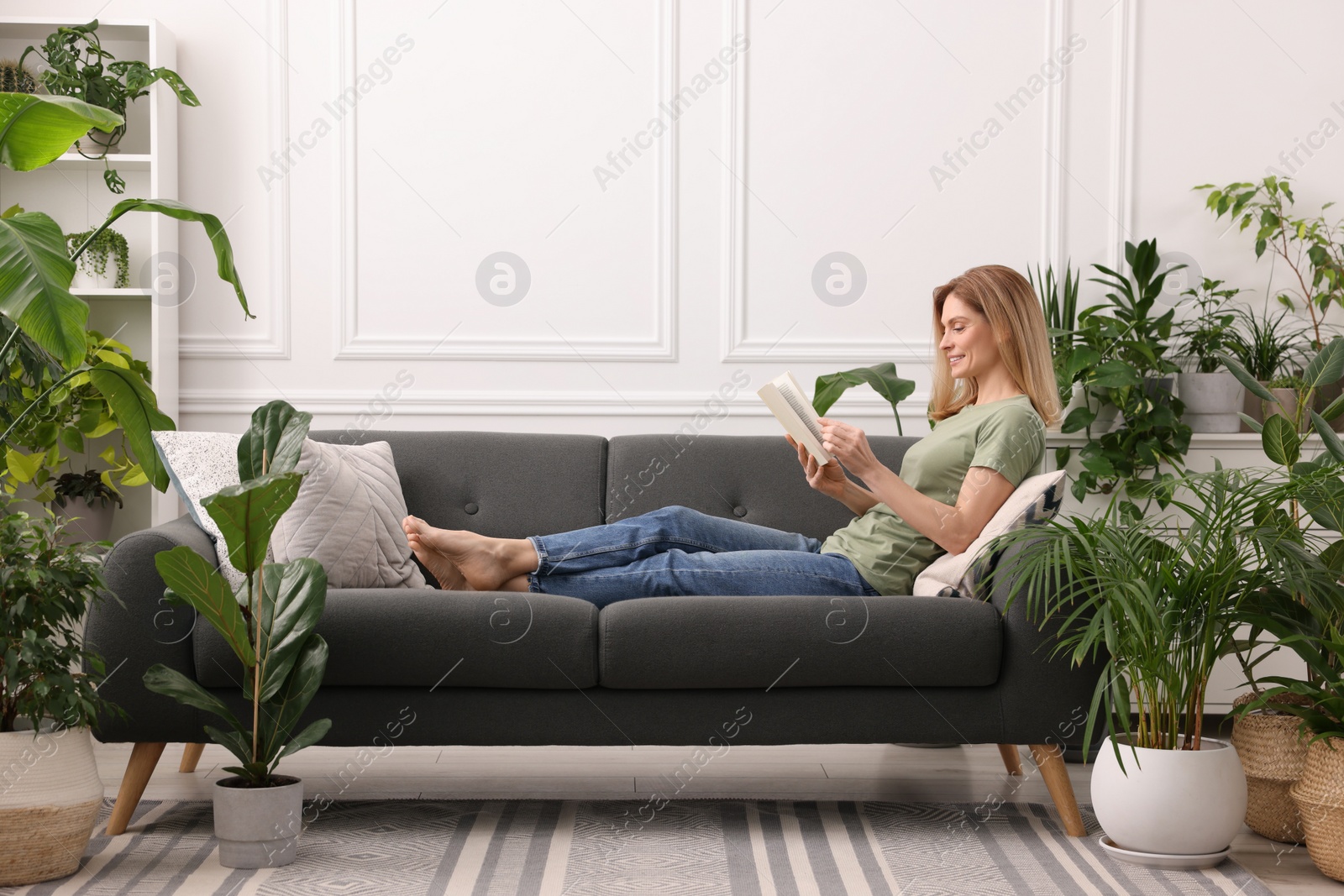 Photo of Woman reading book on sofa surrounded by beautiful potted houseplants at home