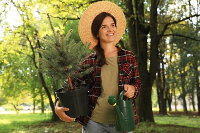 Photo of Young woman holding pot with conifer tree and watering can in park on sunny day