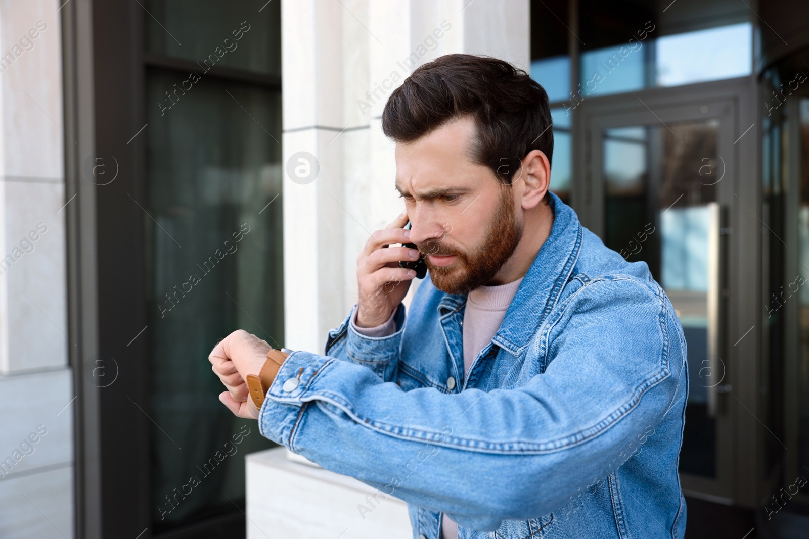 Photo of Emotional man talking on smartphone and looking at watch near building. Being late concept