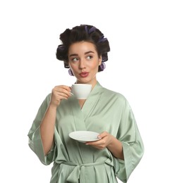 Young woman in silk bathrobe with hair curlers holding cup of drink on white background