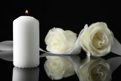 Burning candle, white roses and ribbon on black mirror surface in darkness, closeup with space for text. Funeral symbols