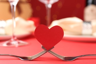 Joined forks with paper heart on red table, closeup. Romantic dinner