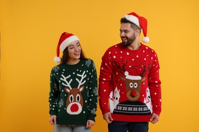 Photo of Happy young couple in Santa hats showing Christmas sweaters on orange background