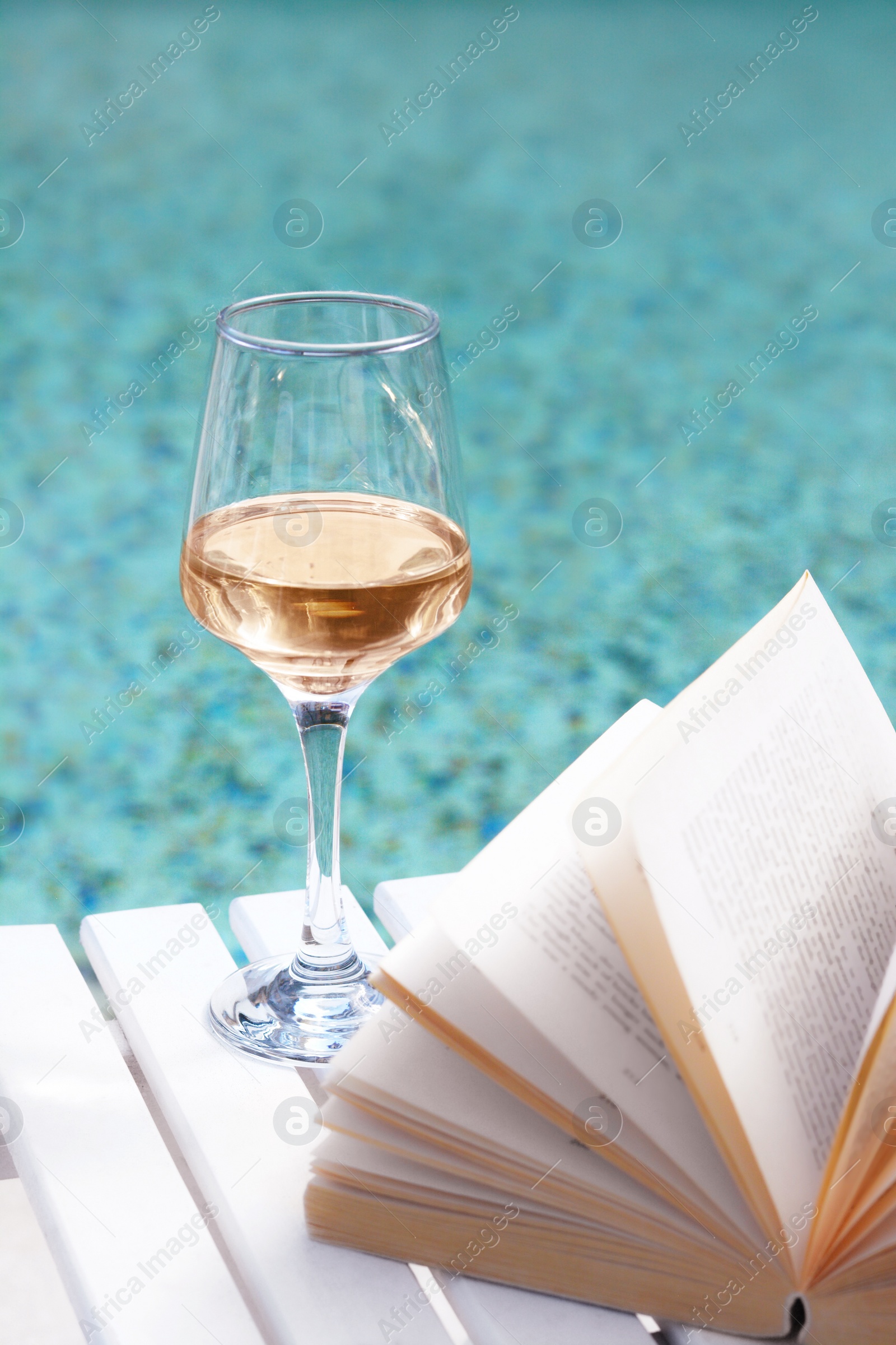 Photo of Glass of tasty wine and open book on wooden table near swimming pool