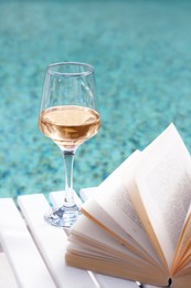Glass of tasty wine and open book on wooden table near swimming pool