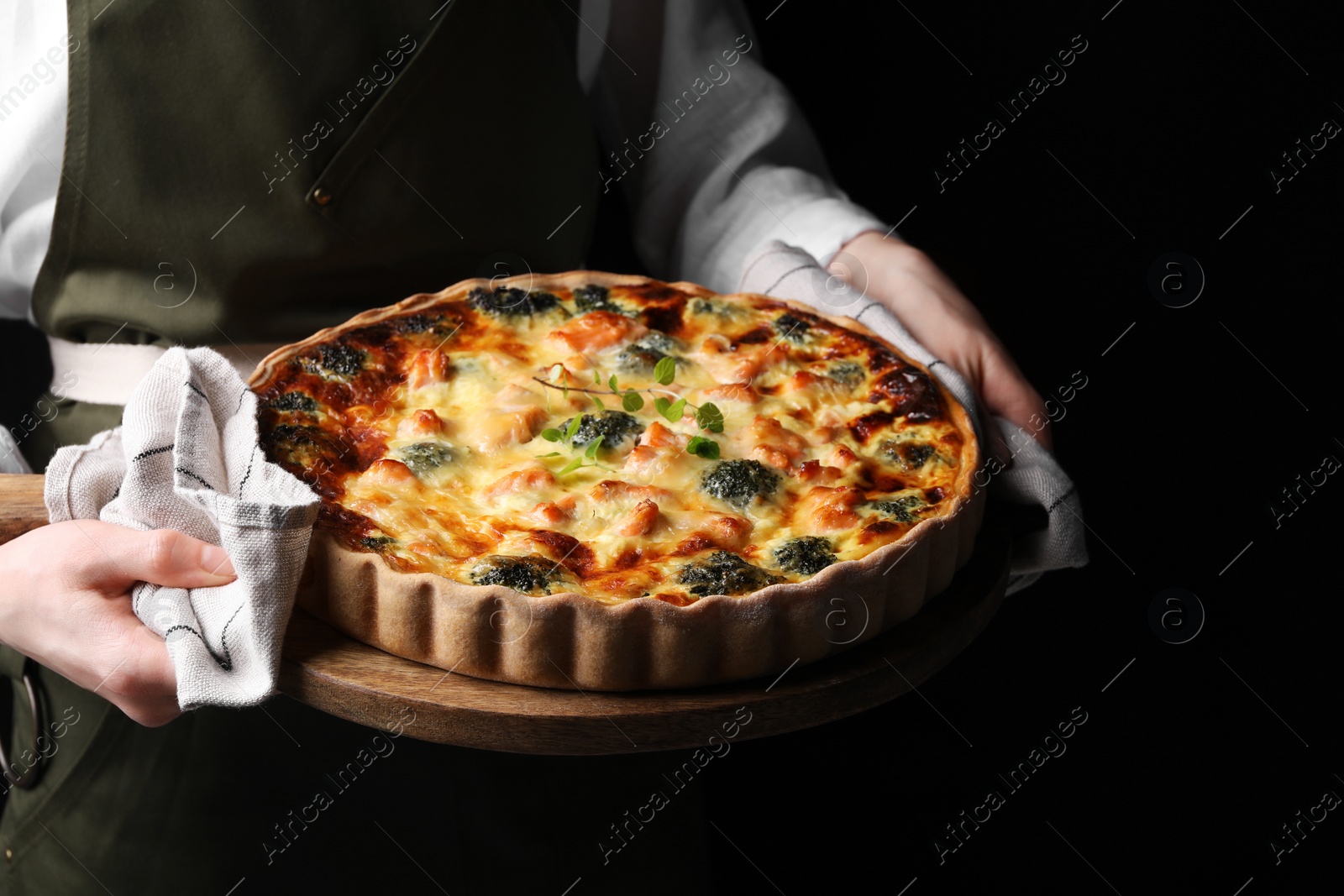 Photo of Woman holding wooden board with delicious homemade salmon quiche on black background, closeup