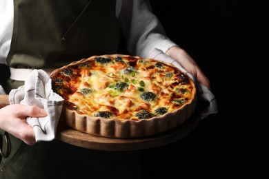 Photo of Woman holding wooden board with delicious homemade salmon quiche on black background, closeup