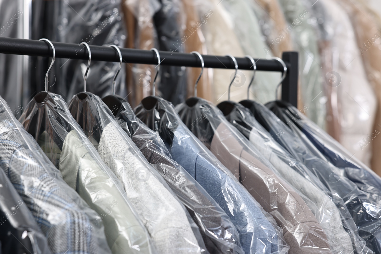 Photo of Dry-cleaning service. Many different clothes in plastic bags hanging on rack indoors, closeup