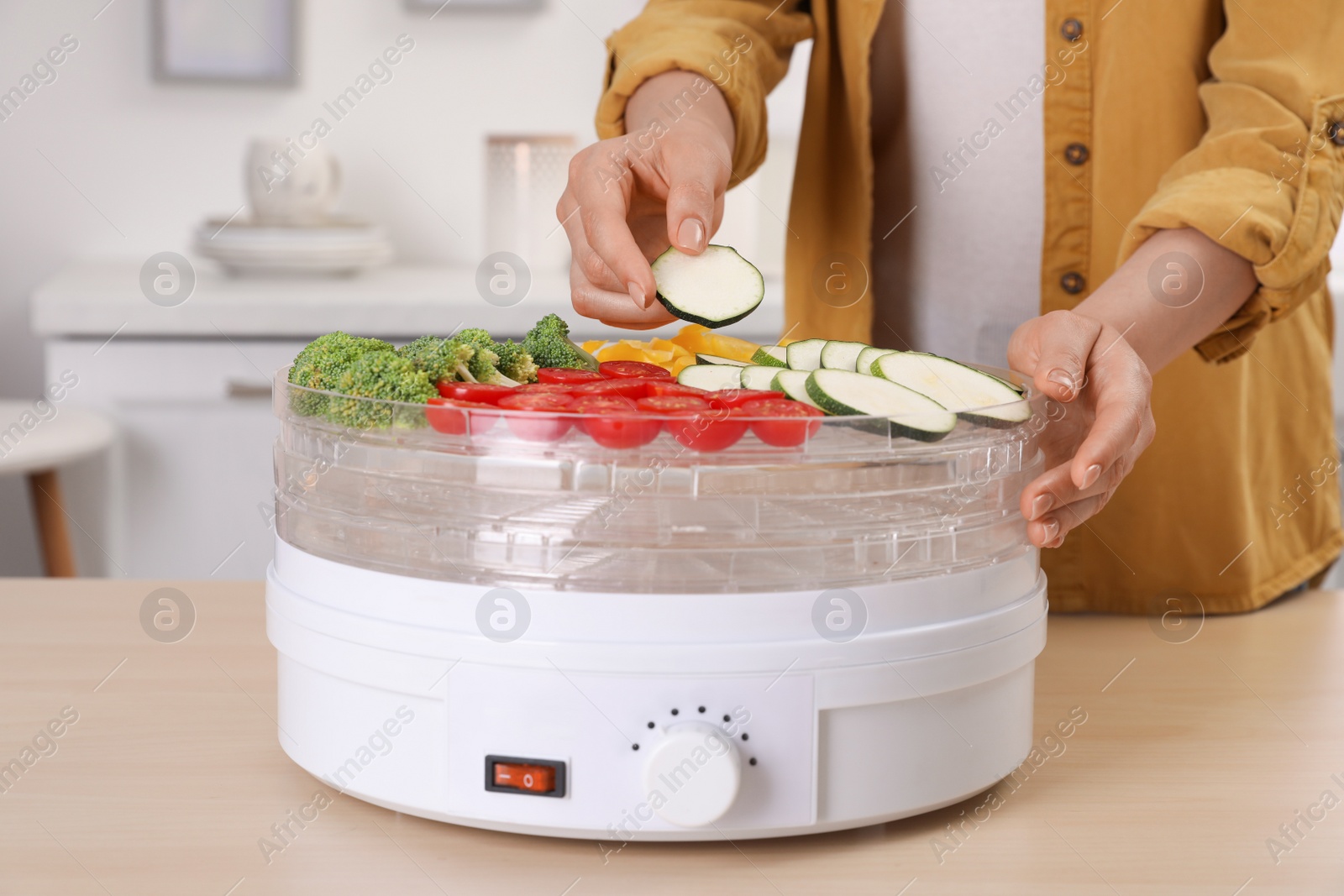 Photo of Woman putting cut zucchini into fruit dehydrator machine at wooden table in kitchen, closeup