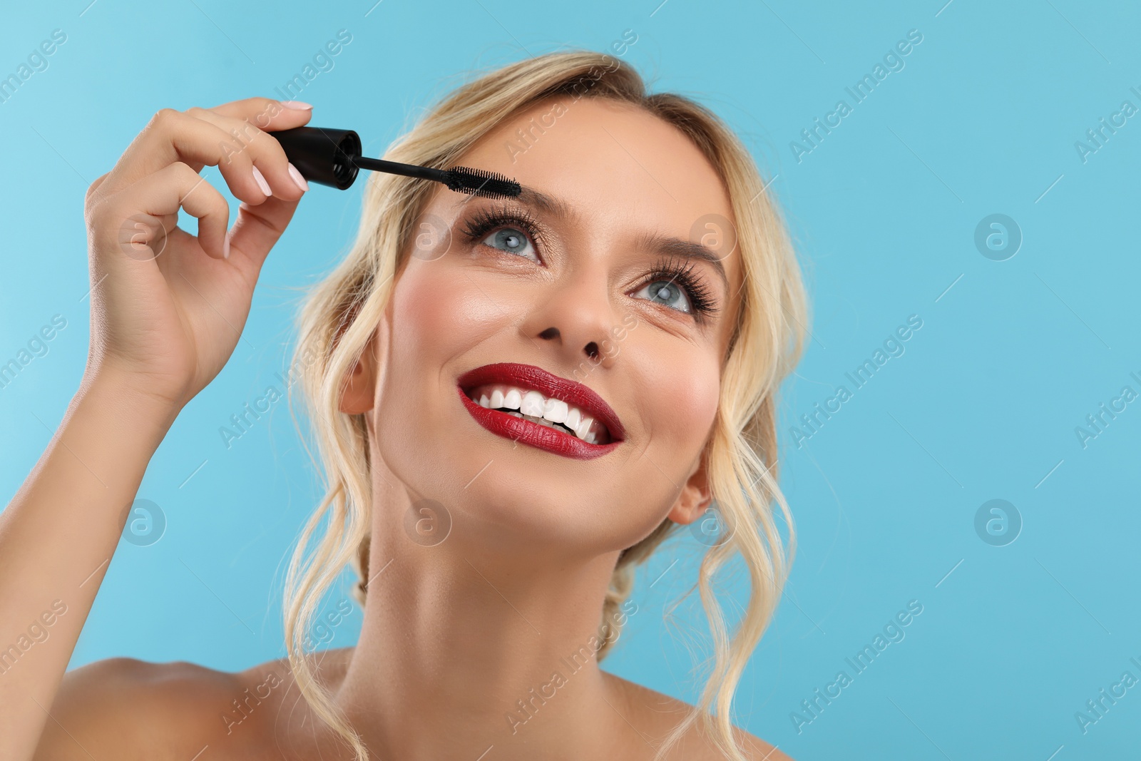 Photo of Beautiful makeup. Smiling woman applying mascara on light blue background