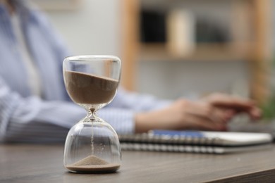 Photo of Hourglass with flowing sand on desk. Woman using laptop indoors, selective focus