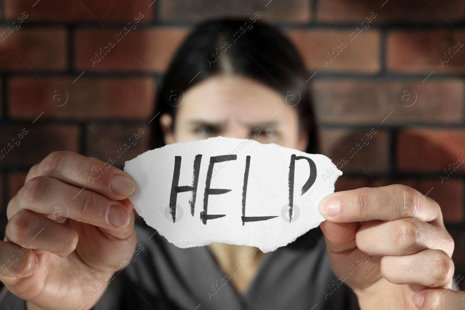 Photo of Domestic violence concept. Unhappy woman holding paper with written word Help near brick wall, focus on hands