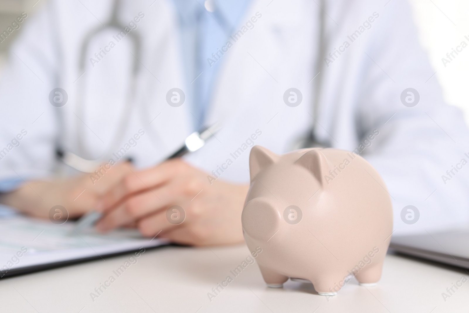 Photo of Doctor making notes at white table, focus on piggy bank